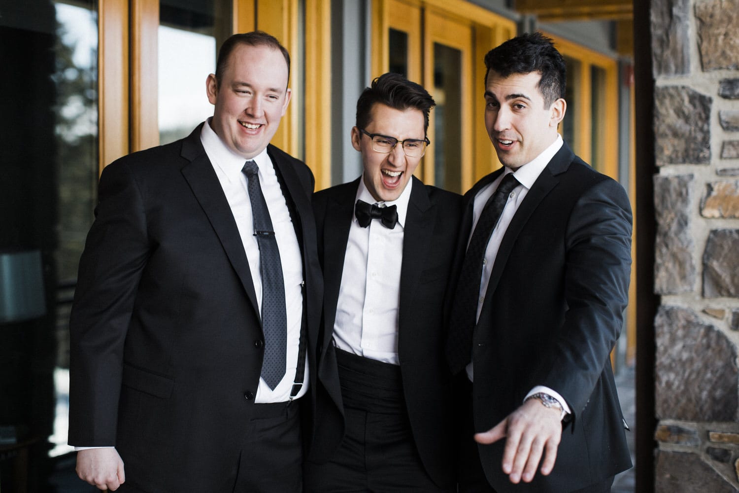 Three men in tuxedos laughing and posing together in a stylish setting with wooden doors in the background.