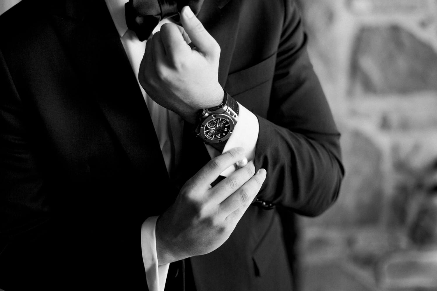 A close-up of a groom's hands adjusting his bow tie while wearing an elegant watch, dressed in a formal suit.