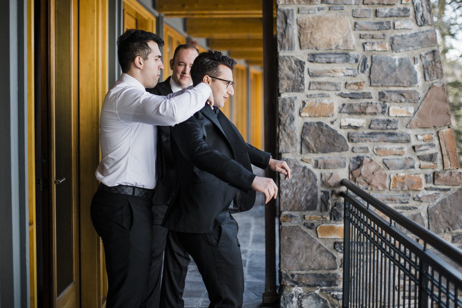 A groom being adjusted by his groomsmen, standing on a balcony with wooden and stone architecture in the background.