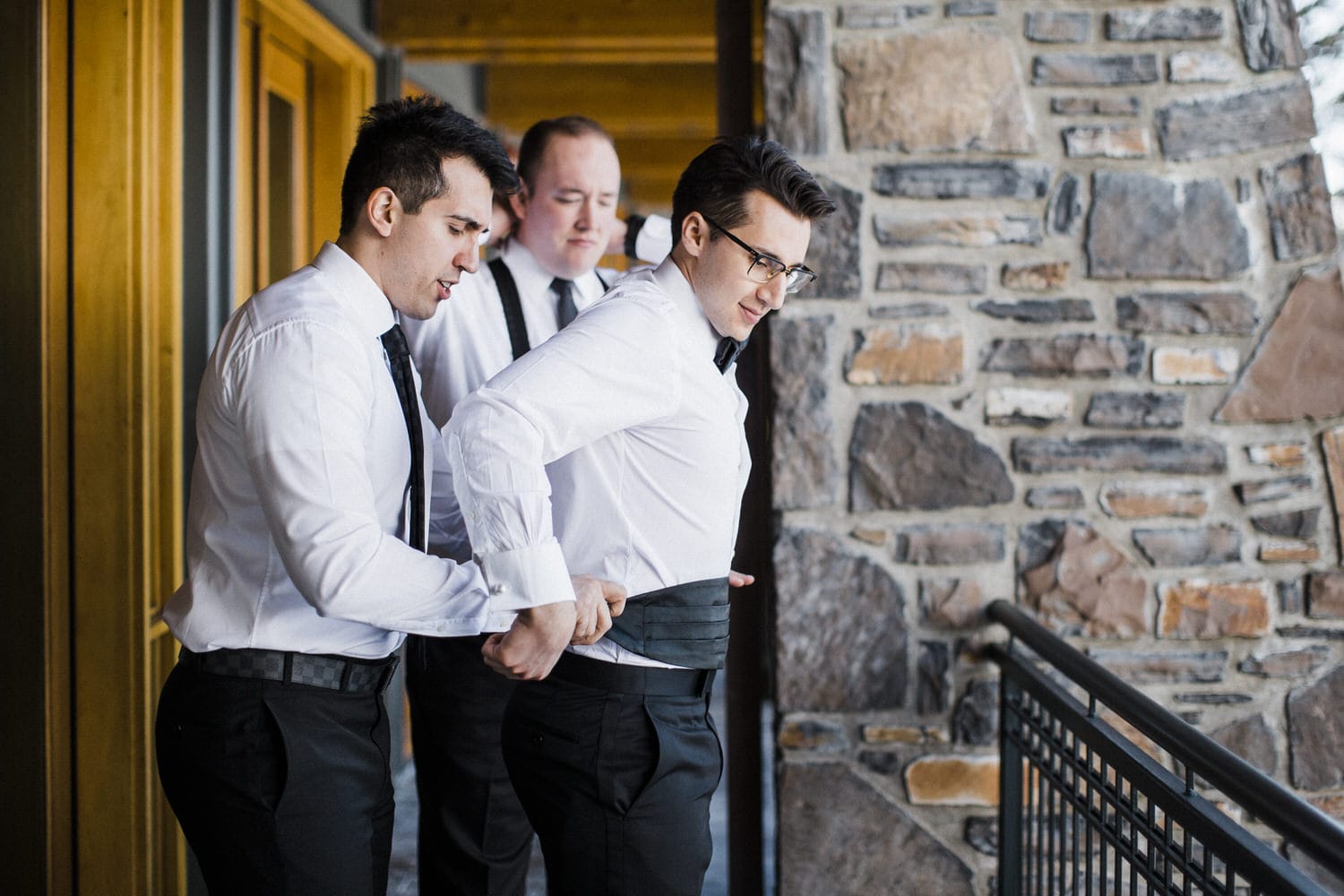 A groom receives assistance from his friends as they prepare for an event, featuring a rustic stone backdrop and elegant attire.