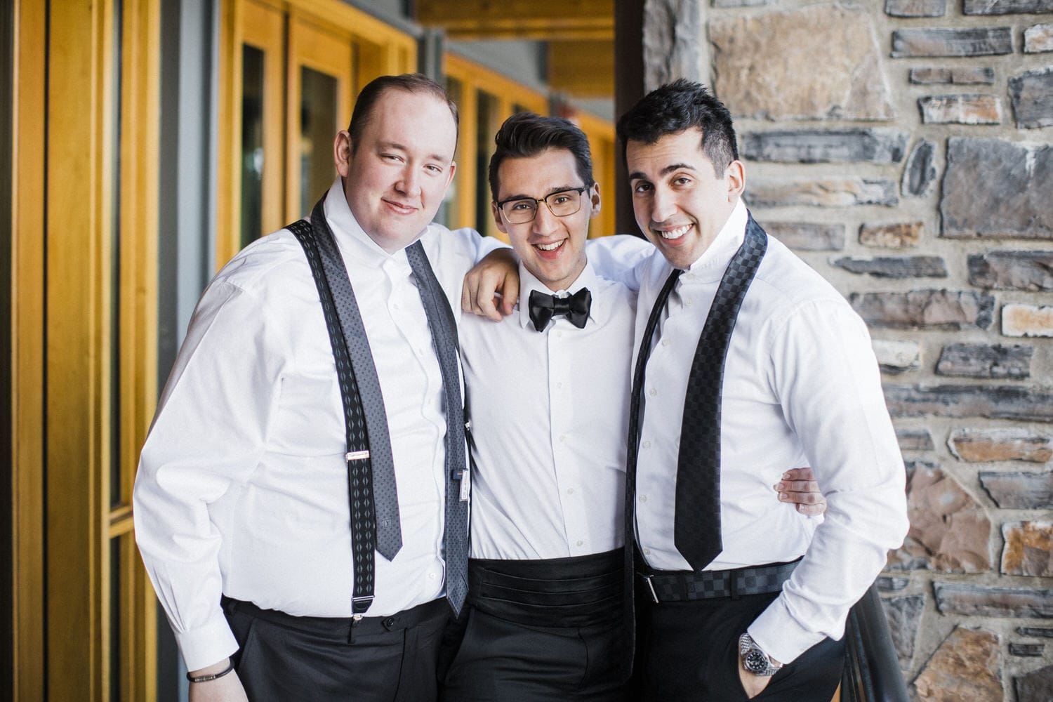 Three well-dressed men posing together in a cheerful moment against a rustic stone wall.