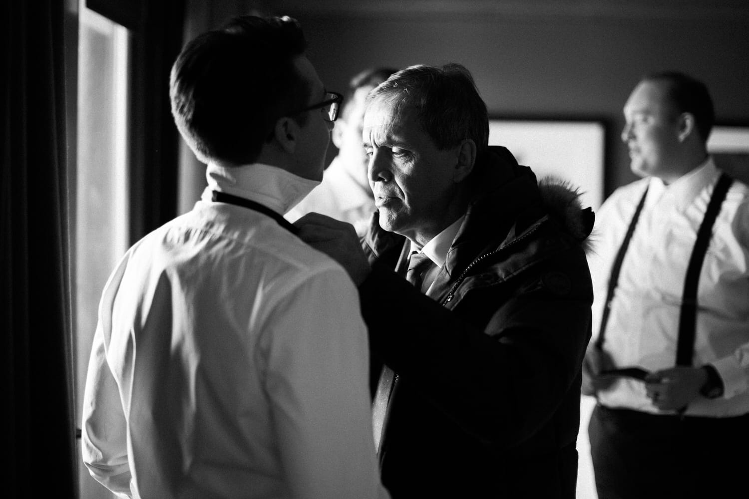 Preparation Moments::A man in a white shirt receiving assistance with his collar while another man looks on in the background, all captured in a soft black and white setting.