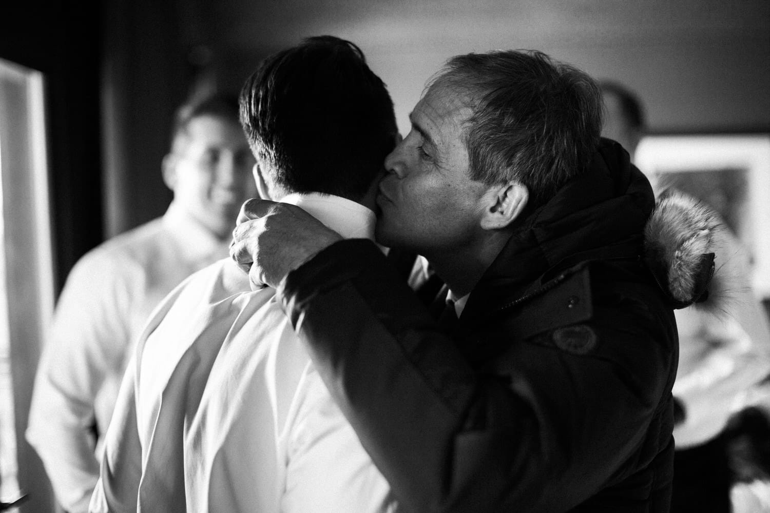 A man lovingly kisses another man on the side of the head, capturing a heartfelt moment before a significant event, with others in the background smiling.