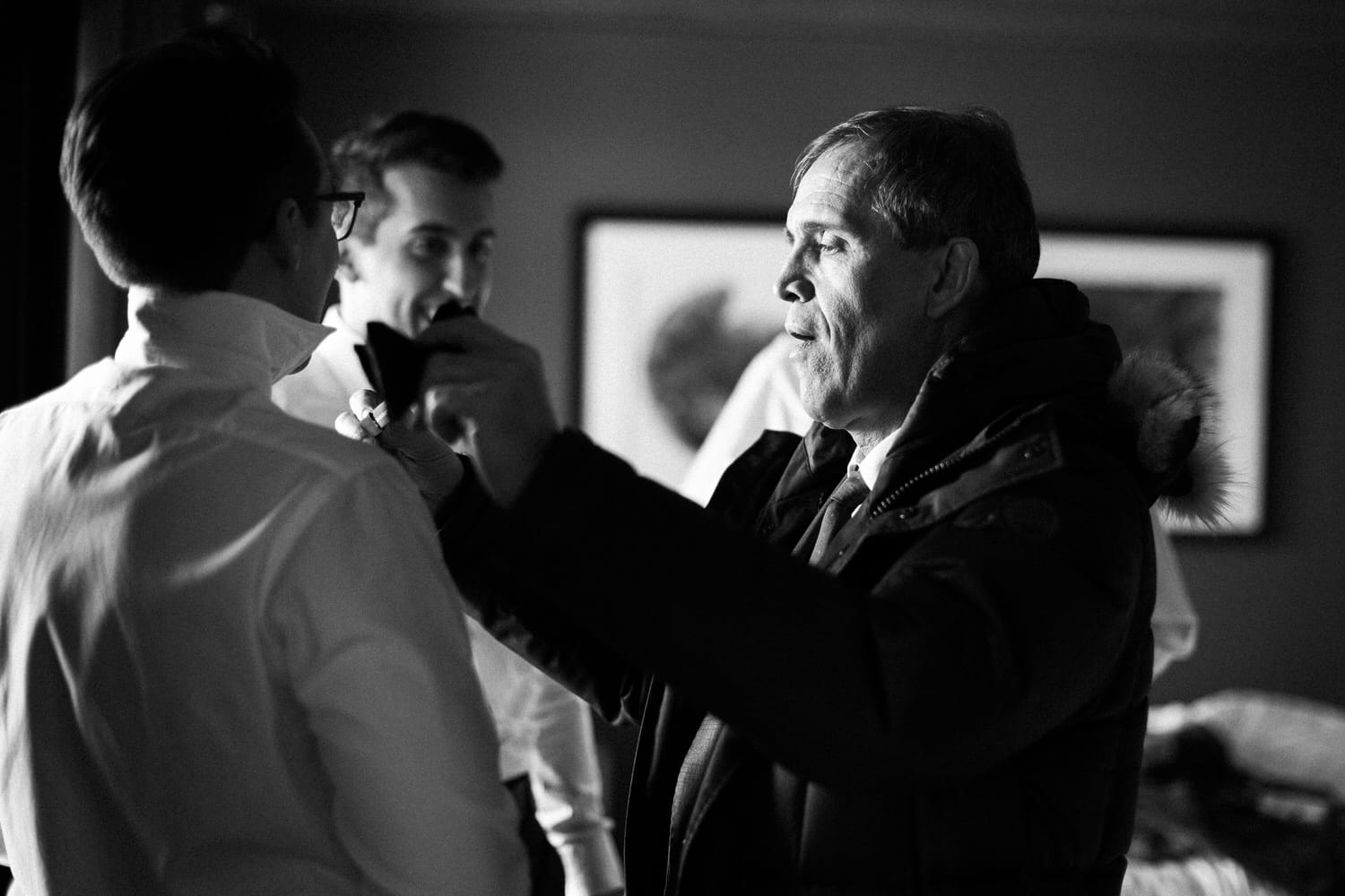 A groom being assisted with his attire while another man smiles in the background, captured in a black and white setting.