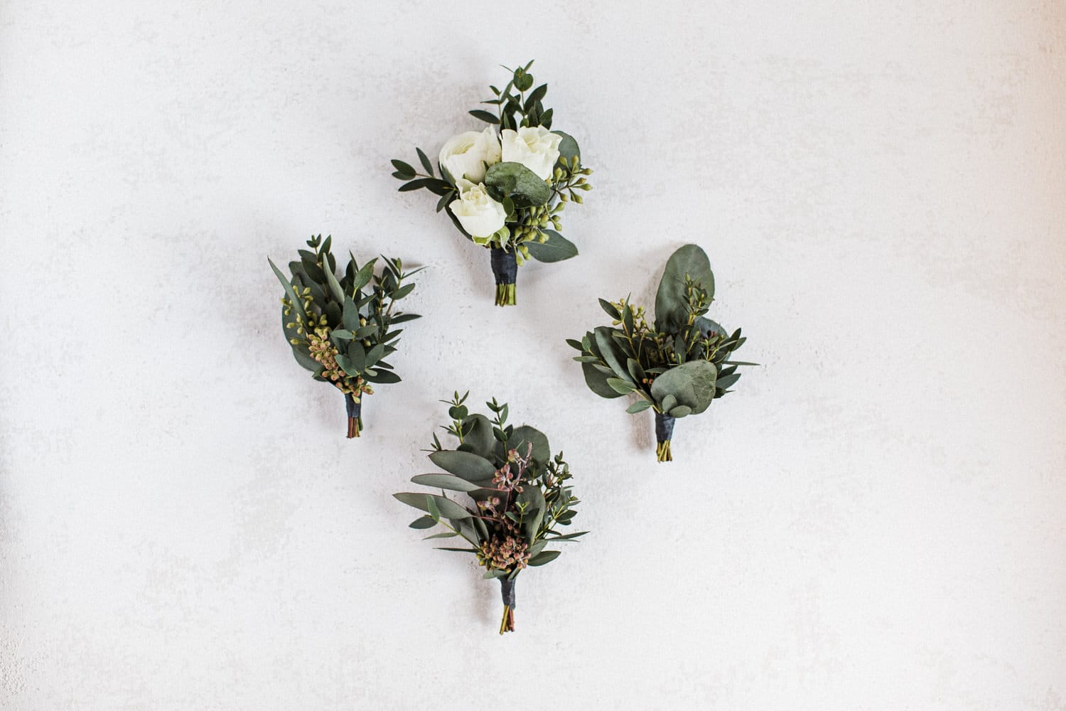 A collection of four elegant floral boutonnieres featuring greenery and white roses arranged against a textured white background.