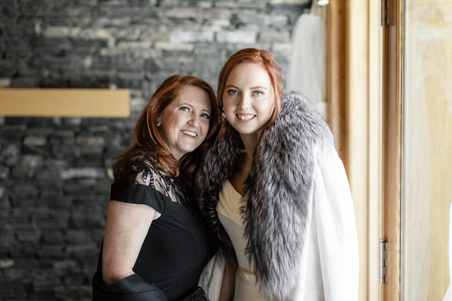 A smiling mother and daughter pose together indoors, showcasing their elegant outfits against a stylish stone wall backdrop.