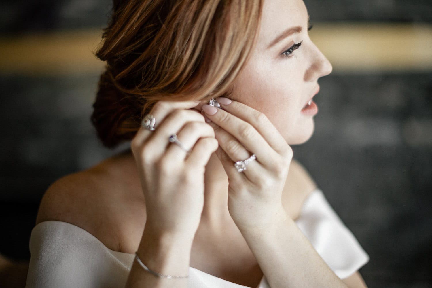 A woman in a bridal gown delicately adjusts her earrings, showcasing her engagement rings and subtle makeup.