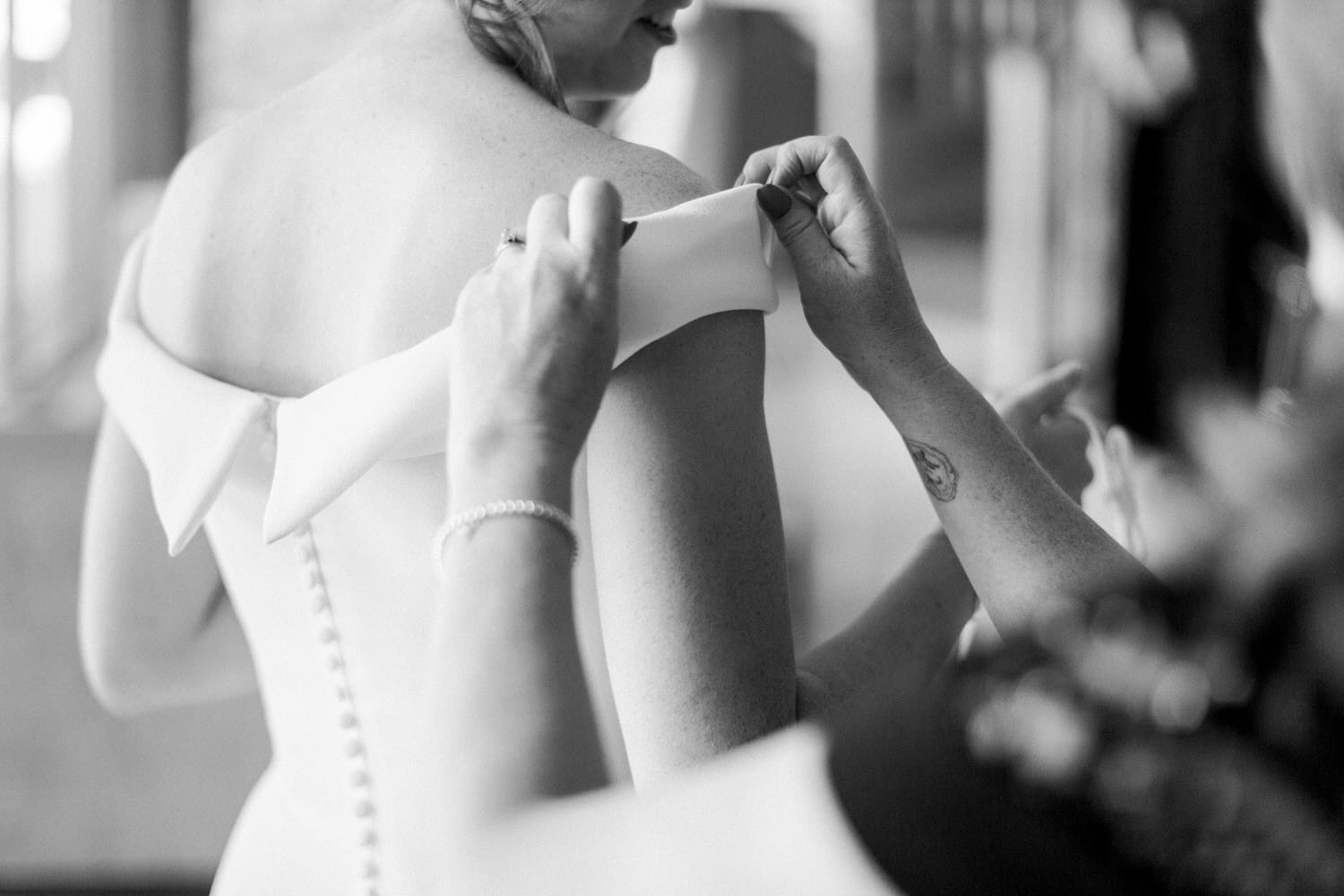 A close-up of a woman helping another adjust the off-shoulder neckline of a wedding dress.