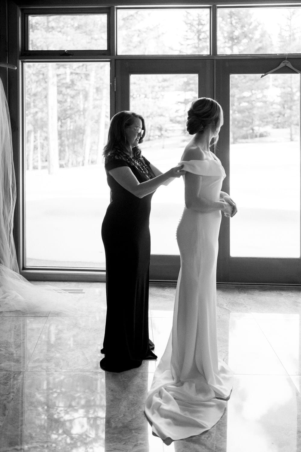 A bride is assisted by a woman in dressing for her wedding, captured in a serene, natural light setting.