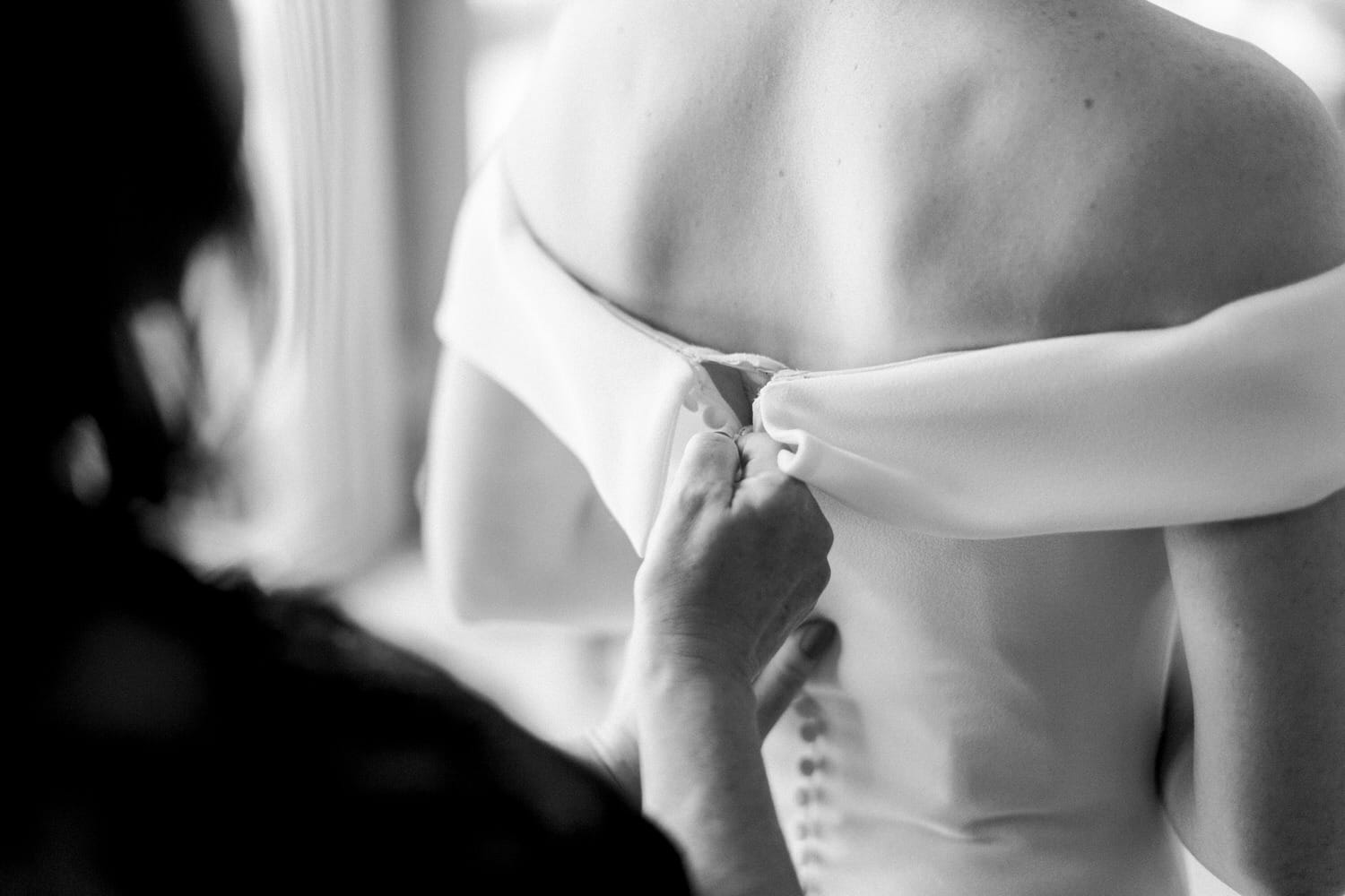 A close-up of a hand fastening the dress of a bride, showcasing the delicate details of the gown and the intimate atmosphere of the moment.