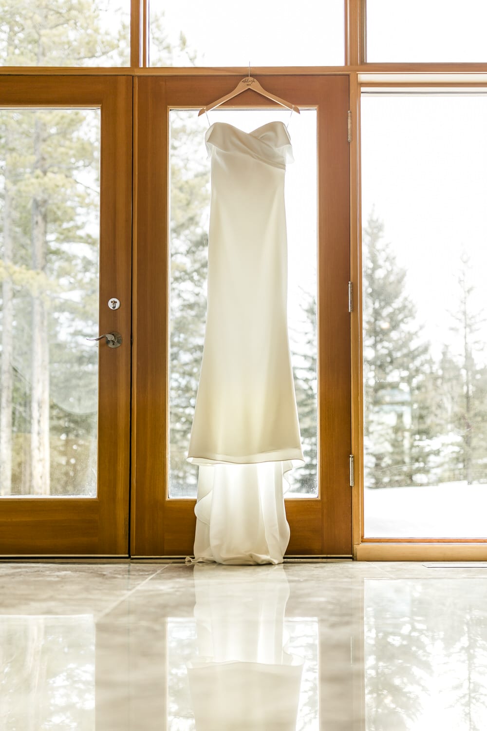 A beautiful white wedding dress hangs gracefully on a hanger in front of large windows, reflecting on a polished floor with a snowy landscape outside.