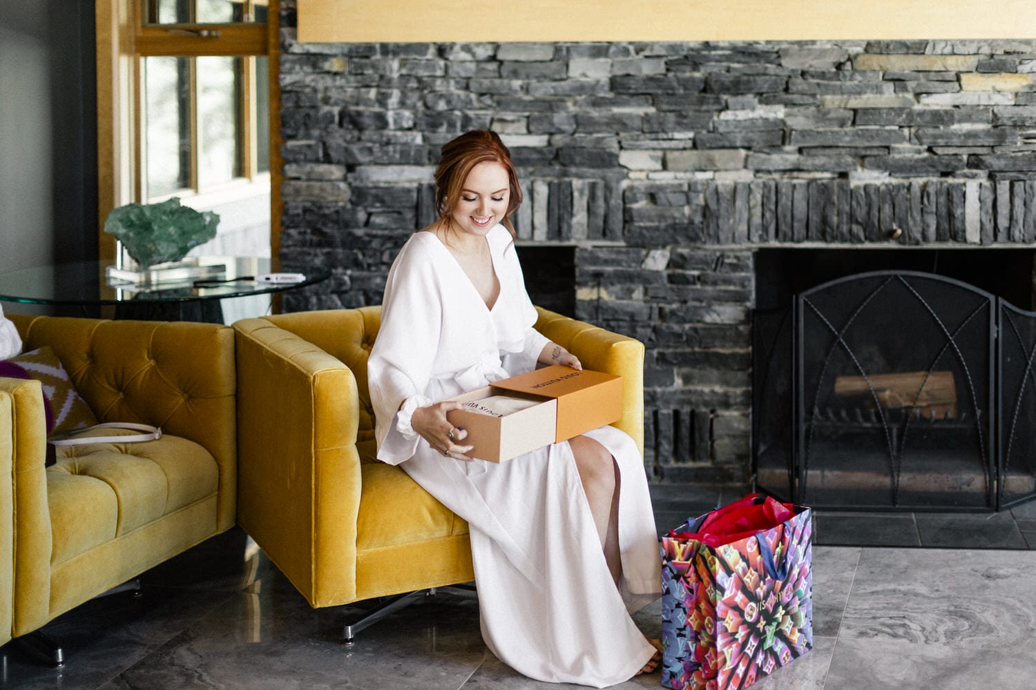 A woman in a white dress smiles while holding two gift boxes, sitting on a yellow sofa in a stylish living room with a stone wall and a colorful shopping bag nearby.