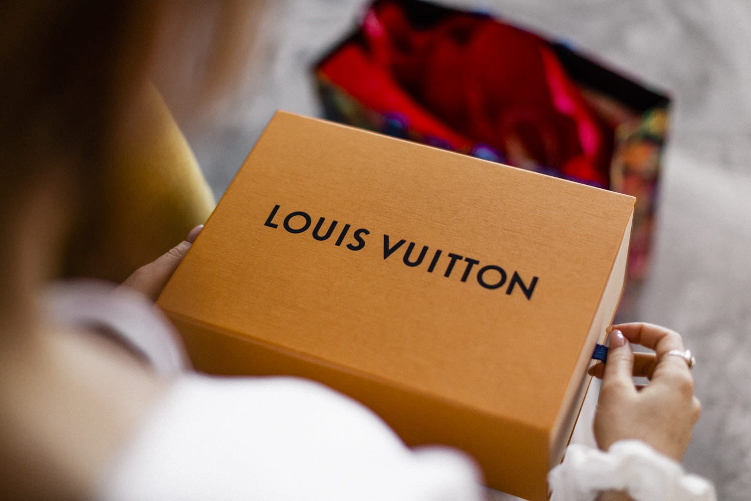 An individual holding a Louis Vuitton box, with a colorful shopping bag visible in the background.