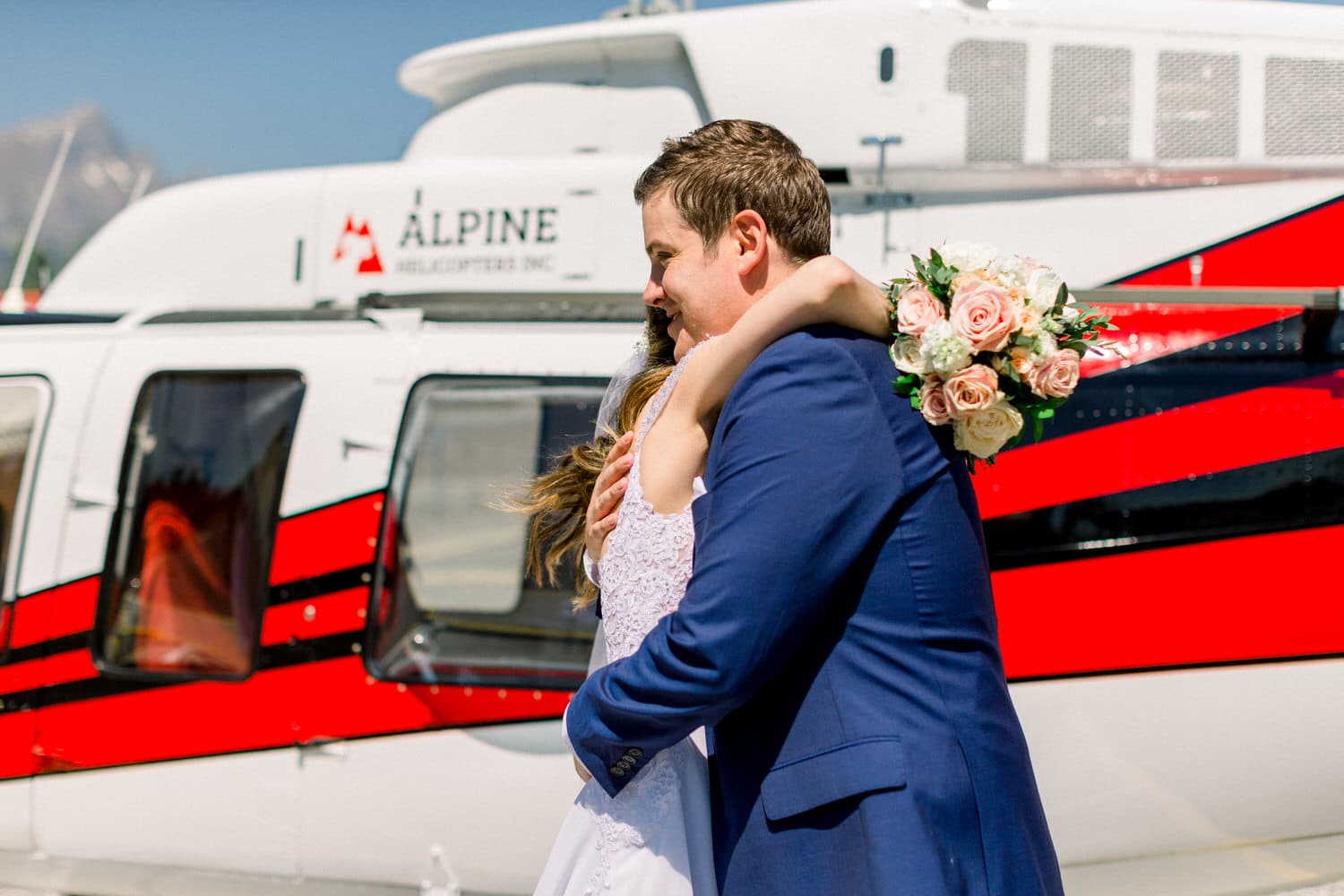 A couple shares a heartfelt hug in front of a helicopter, with the bride holding a bouquet and wearing a white dress, as the groom dons a blue suit.