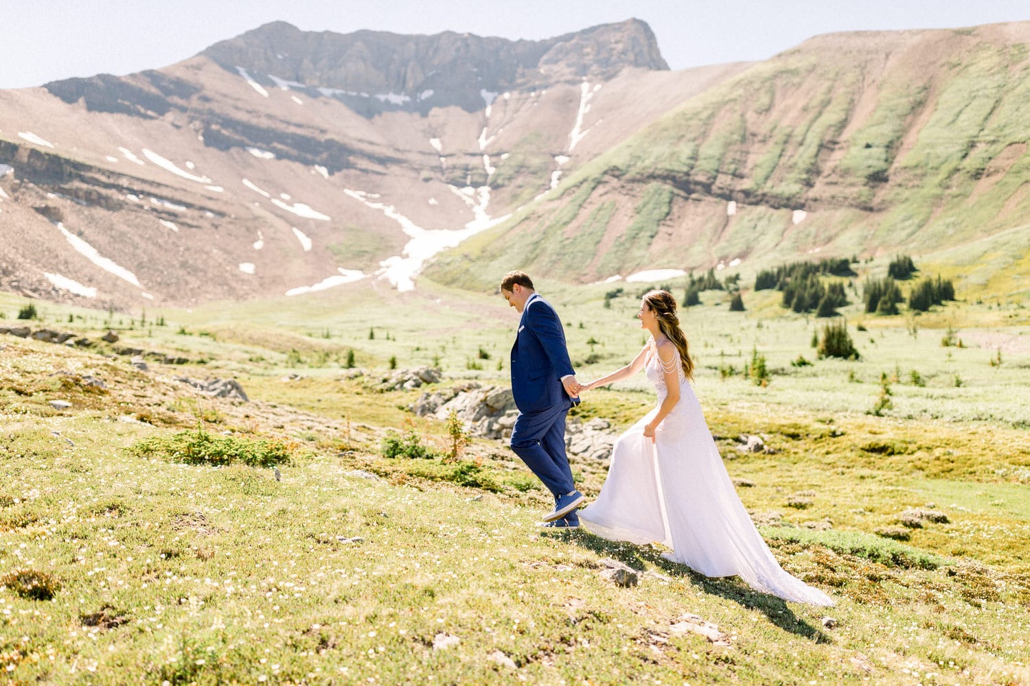 A couple walks hand-in-hand through a lush green landscape with mountains in the background, celebrating their joyful moment together.