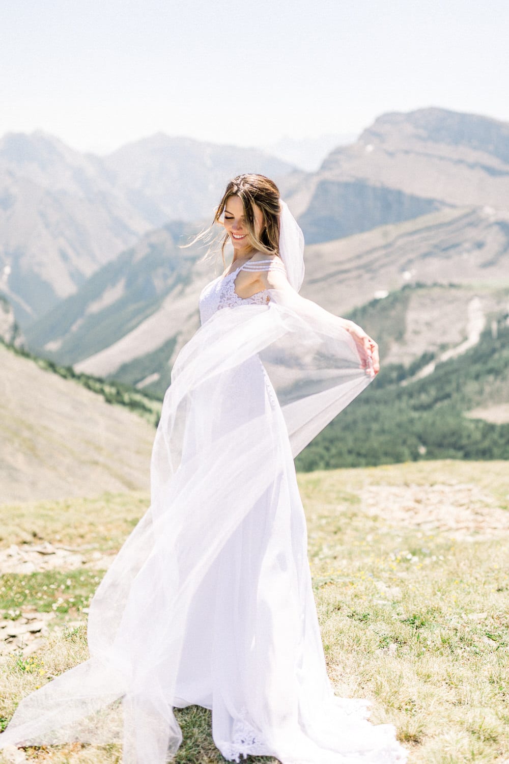 A bride in a flowing white dress and veil gracefully twirls on a mountainside with a stunning landscape in the background.