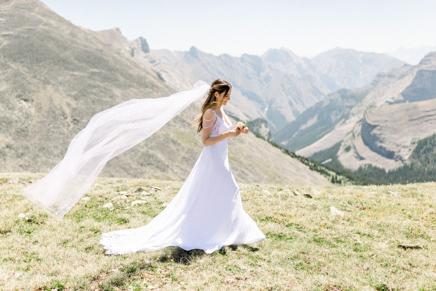 A bride in a flowing white gown walks gracefully on a hillside, with a veil billowing in the wind amidst stunning mountain scenery.