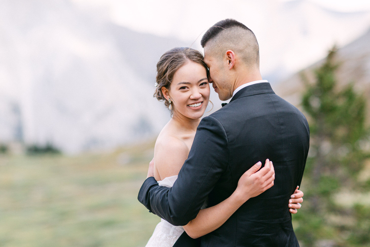 A joyful couple embracing in a scenic outdoor setting, wearing wedding attire, with a backdrop of mountains and greenery.