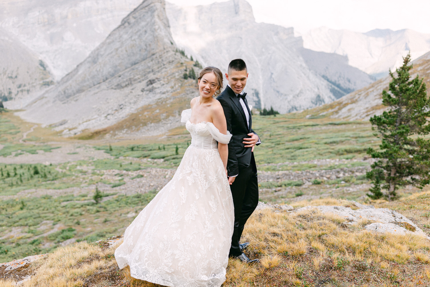 A joyful couple poses in a stunning mountain landscape, showcasing the bride's elegant gown and the groom's dapper tuxedo.