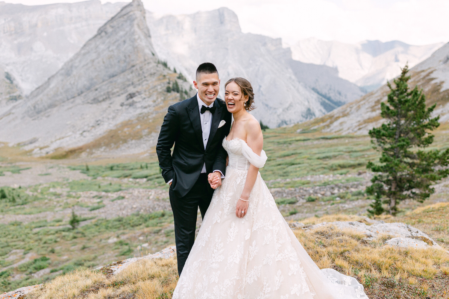 A smiling couple holding hands in a picturesque mountainous landscape, with the bride in a beautiful lace gown and the groom in a classic tuxedo.