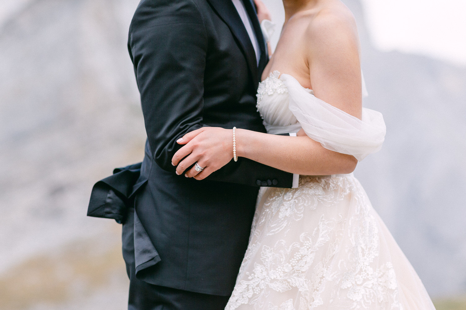 A close-up of a bride and groom embracing, showcasing their elegant attire and wedding rings against a natural backdrop.