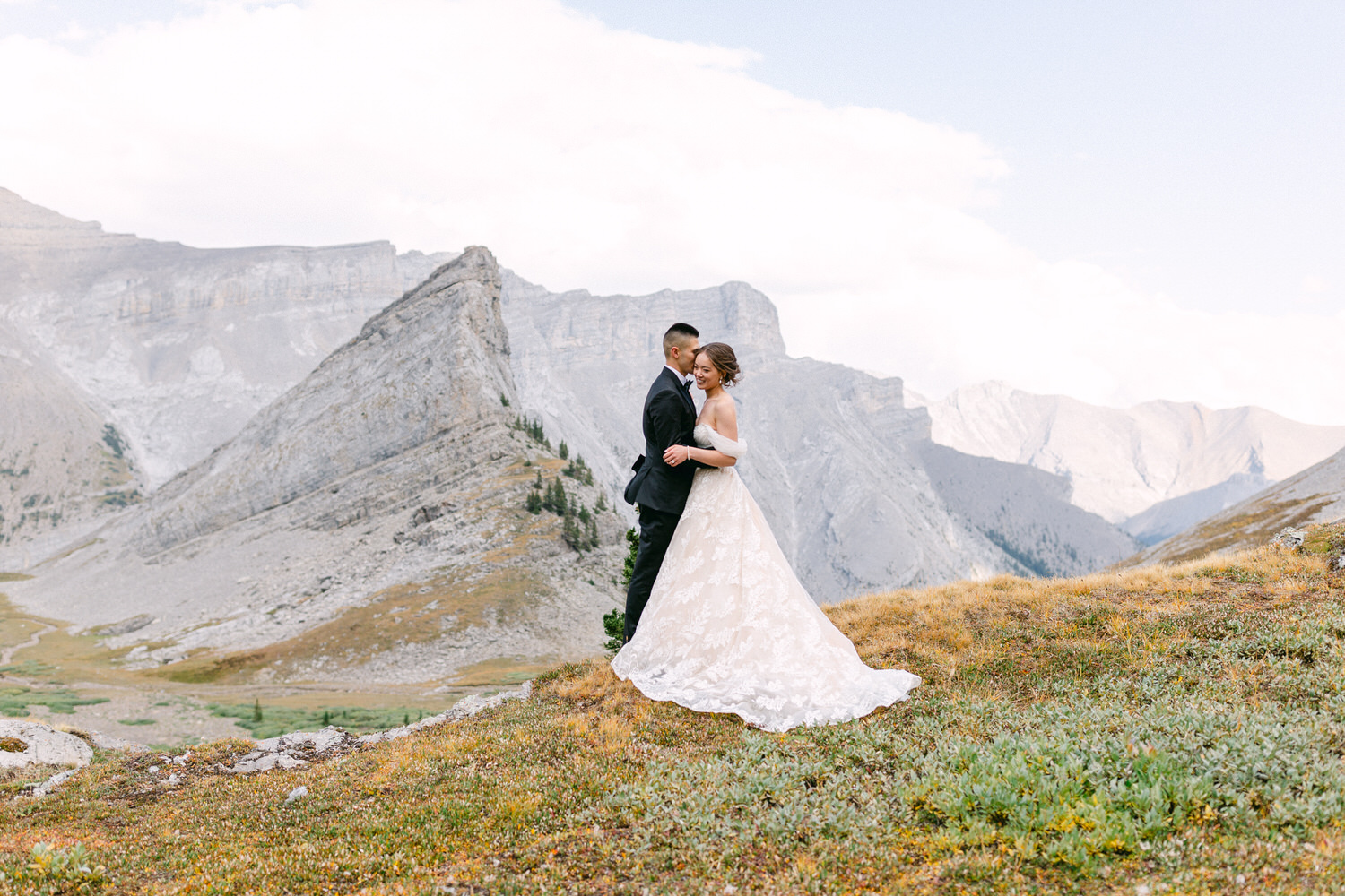 A couple embraces in a scenic mountain landscape, with rolling hills and peaks in the background. The bride wears a beautiful lace gown, while the groom is in a classic suit.