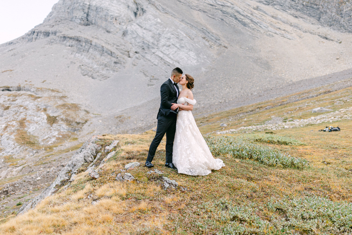 A couple dressed in wedding attire shares a kiss in a picturesque mountainous setting, surrounded by nature and rocky terrain.