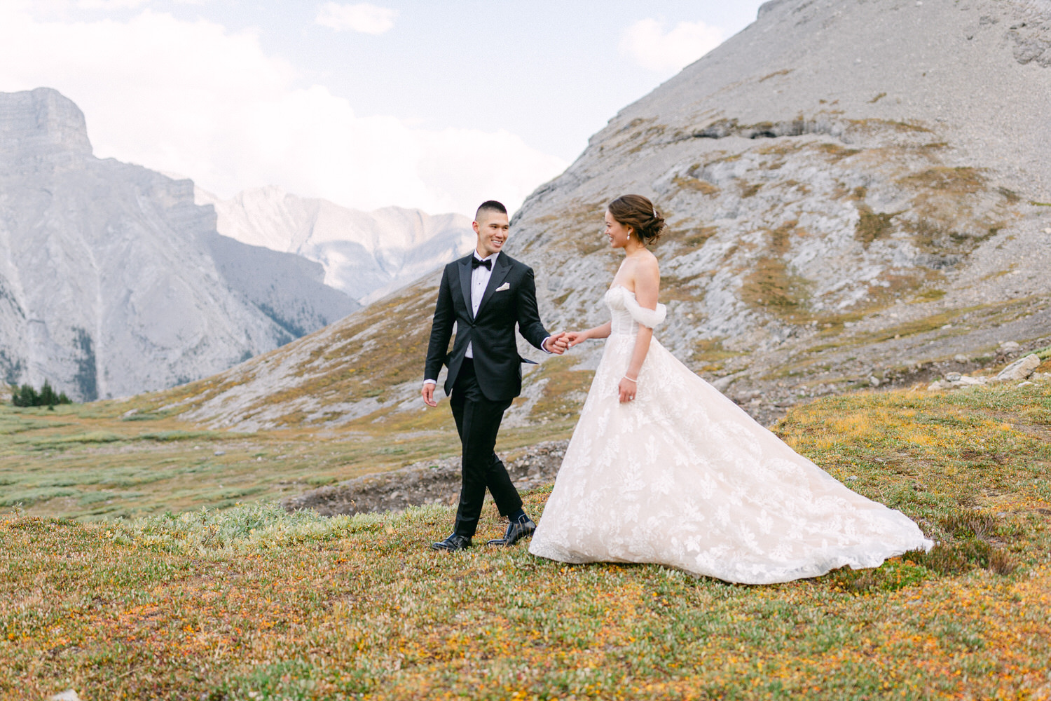 A joyful couple in wedding attire walking hand in hand through a scenic mountainous landscape.