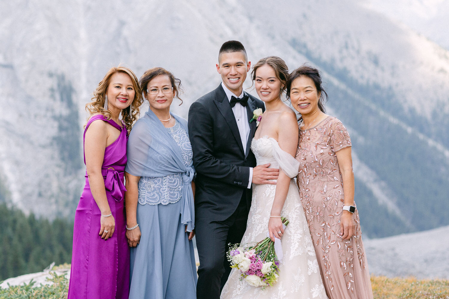 A joyful group portrait of the bride, groom, and four women, elegantly dressed, with a stunning mountain backdrop during a wedding celebration.