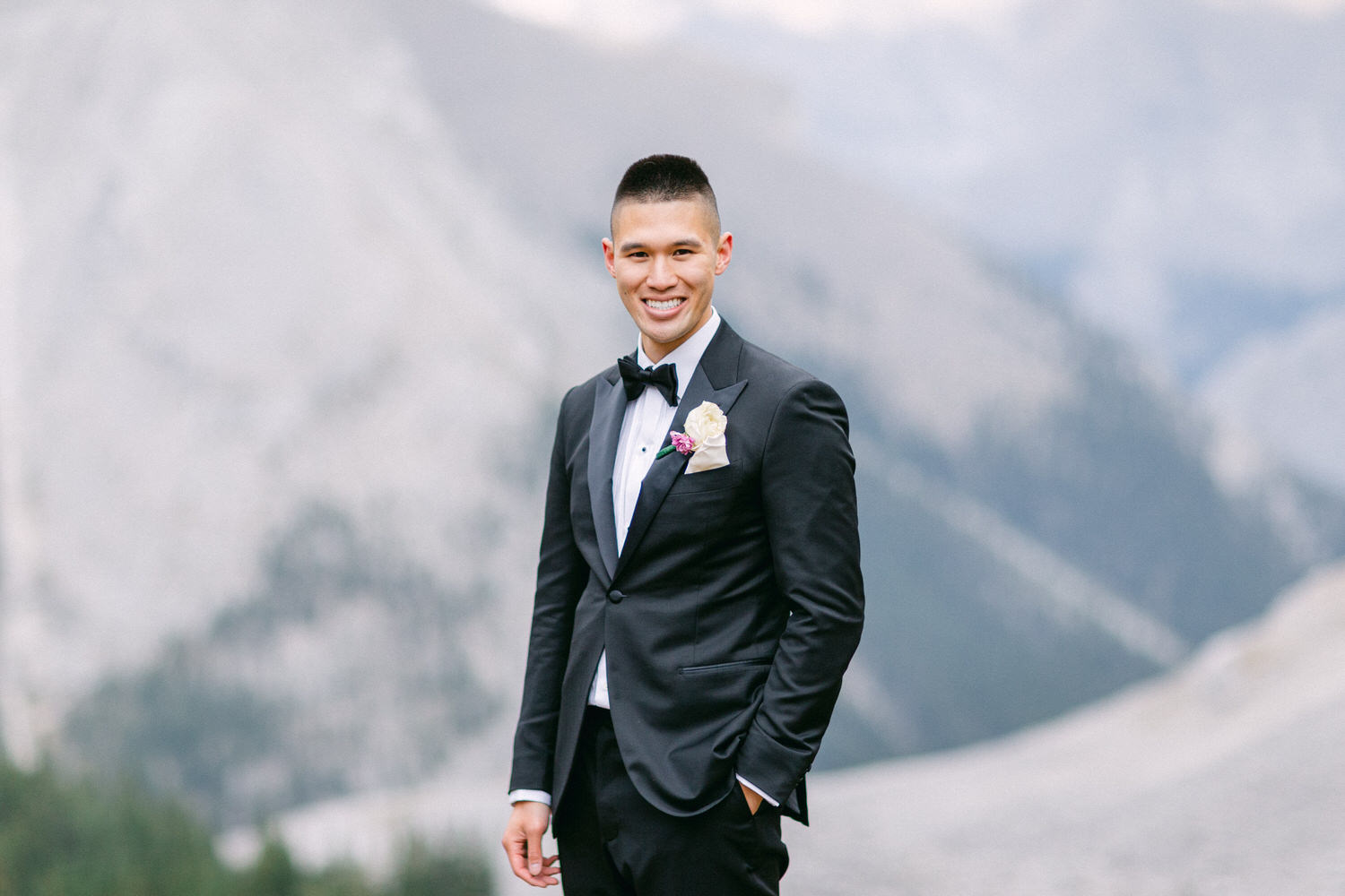 A smiling groom in a tuxedo stands confidently against a majestic mountain backdrop, showcasing natural beauty and elegance.