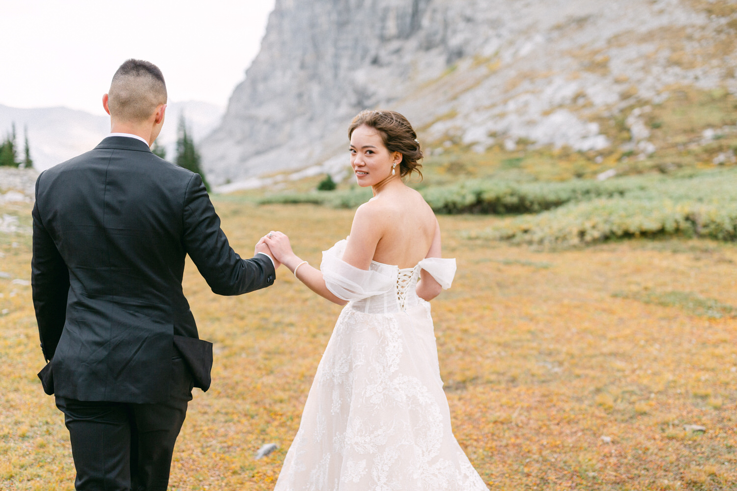 A bride and groom walking hand-in-hand through a scenic landscape, surrounded by mountains and autumn foliage.