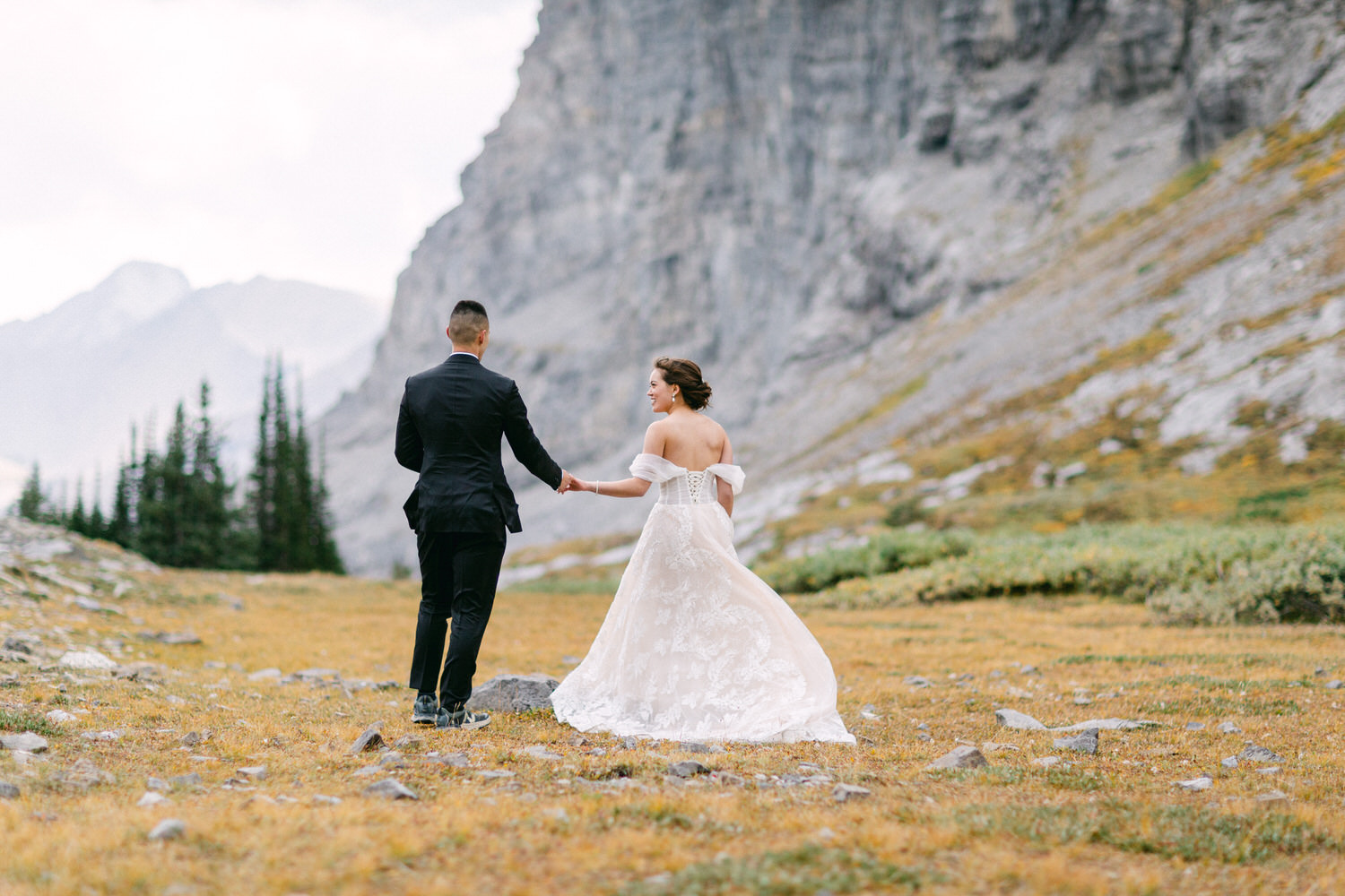 A couple in formal attire walking hand in hand through a picturesque mountain landscape, surrounded by nature and rocky terrain.