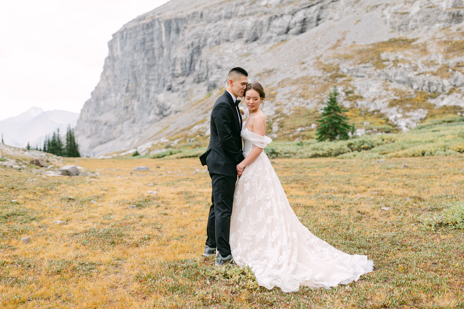 A bride and groom share a tender moment in a scenic mountain landscape, surrounded by vibrant autumn foliage and rocky cliffs.