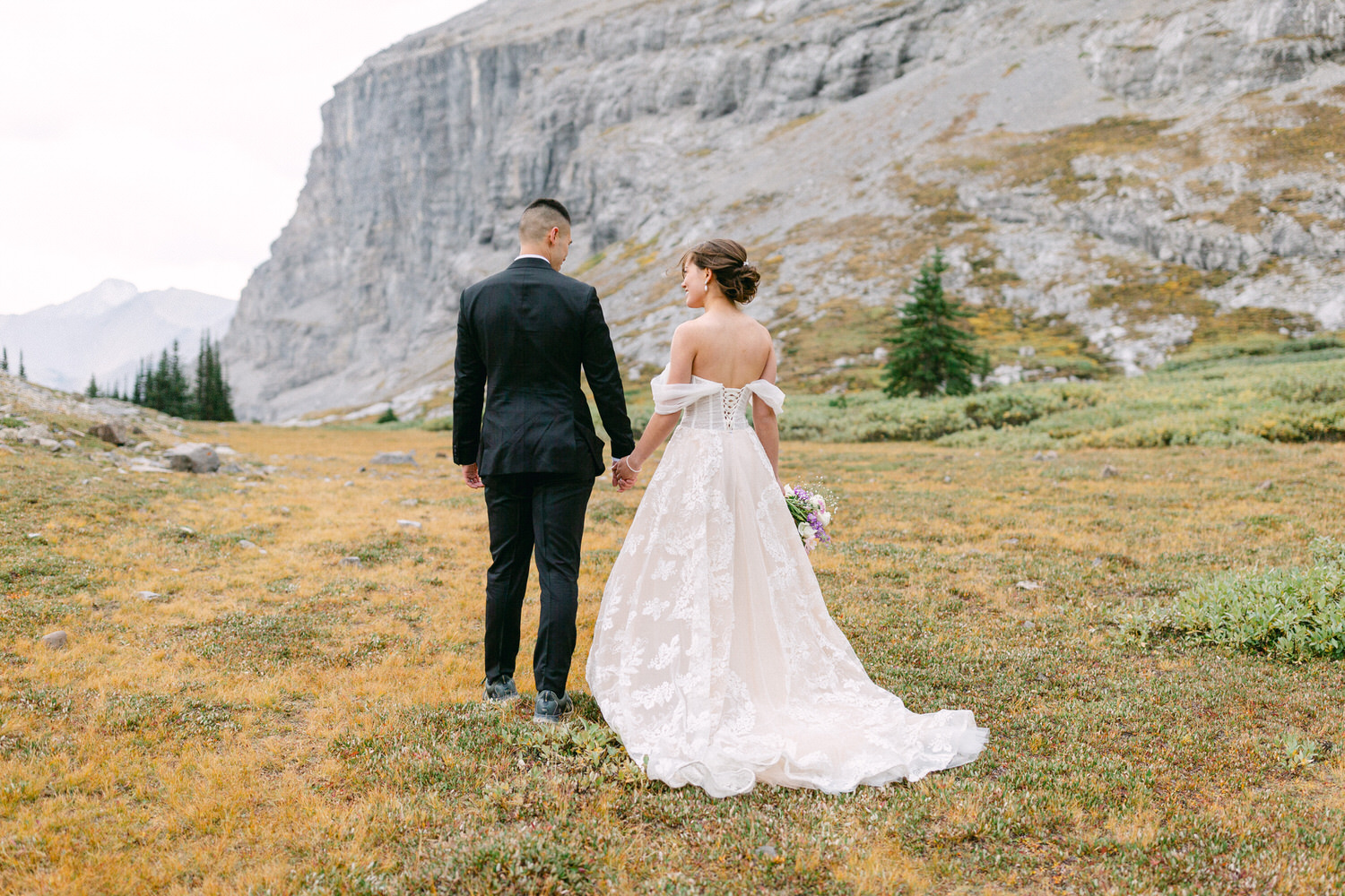 A couple holding hands in an open meadow surrounded by mountain scenery, with the bride in a beautiful dress and holding a bouquet.