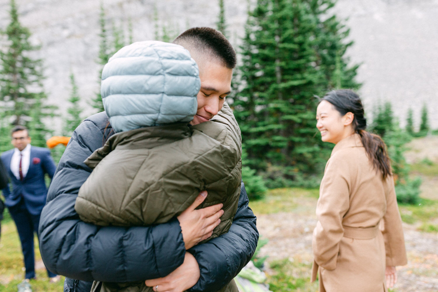 Two individuals share a heartfelt hug in a forested setting, while a third person smiles in the background, showcasing a moment of connection amidst the greenery.