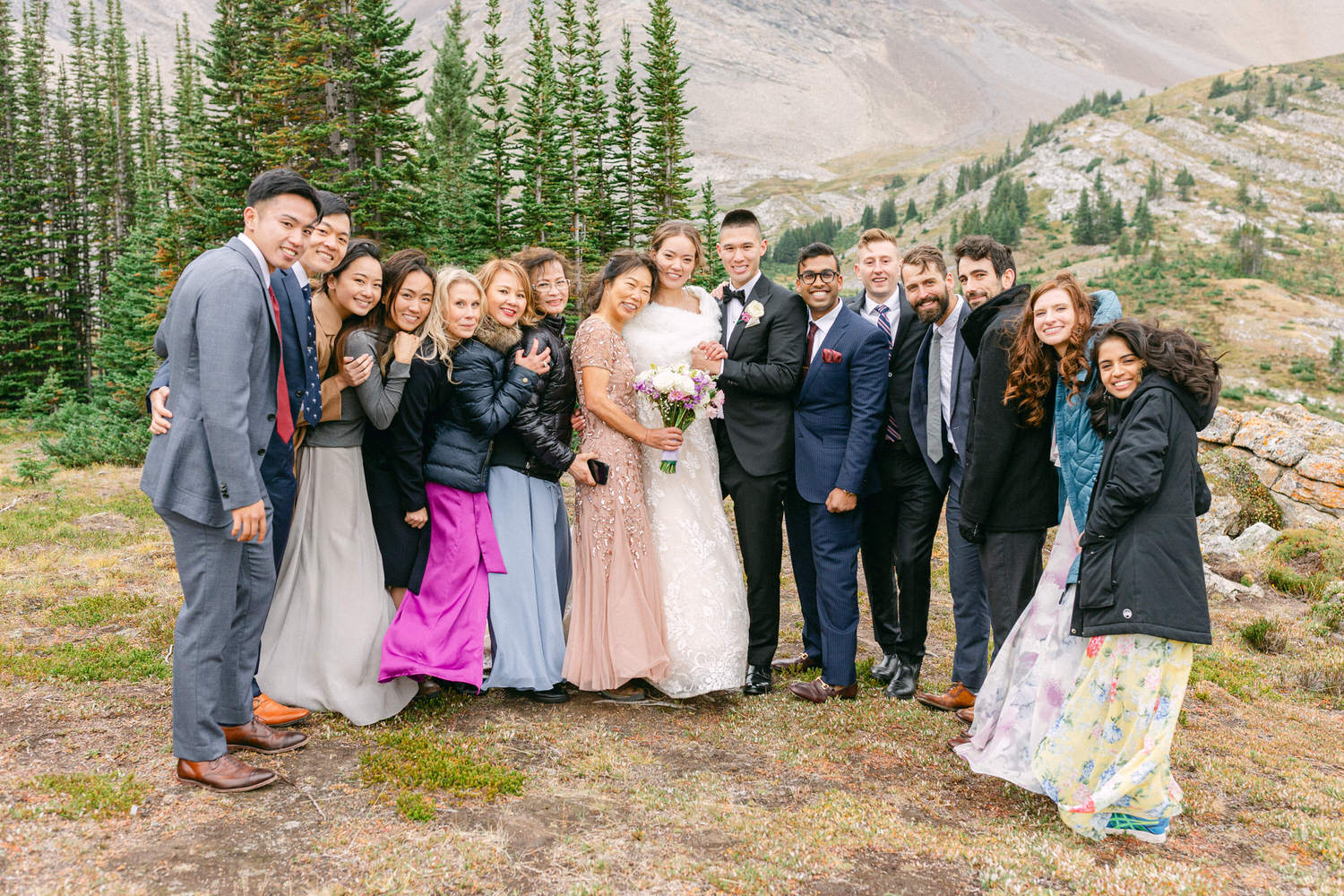 A joyful group photo of wedding guests posing together in a picturesque outdoor setting with mountains and greenery in the background.