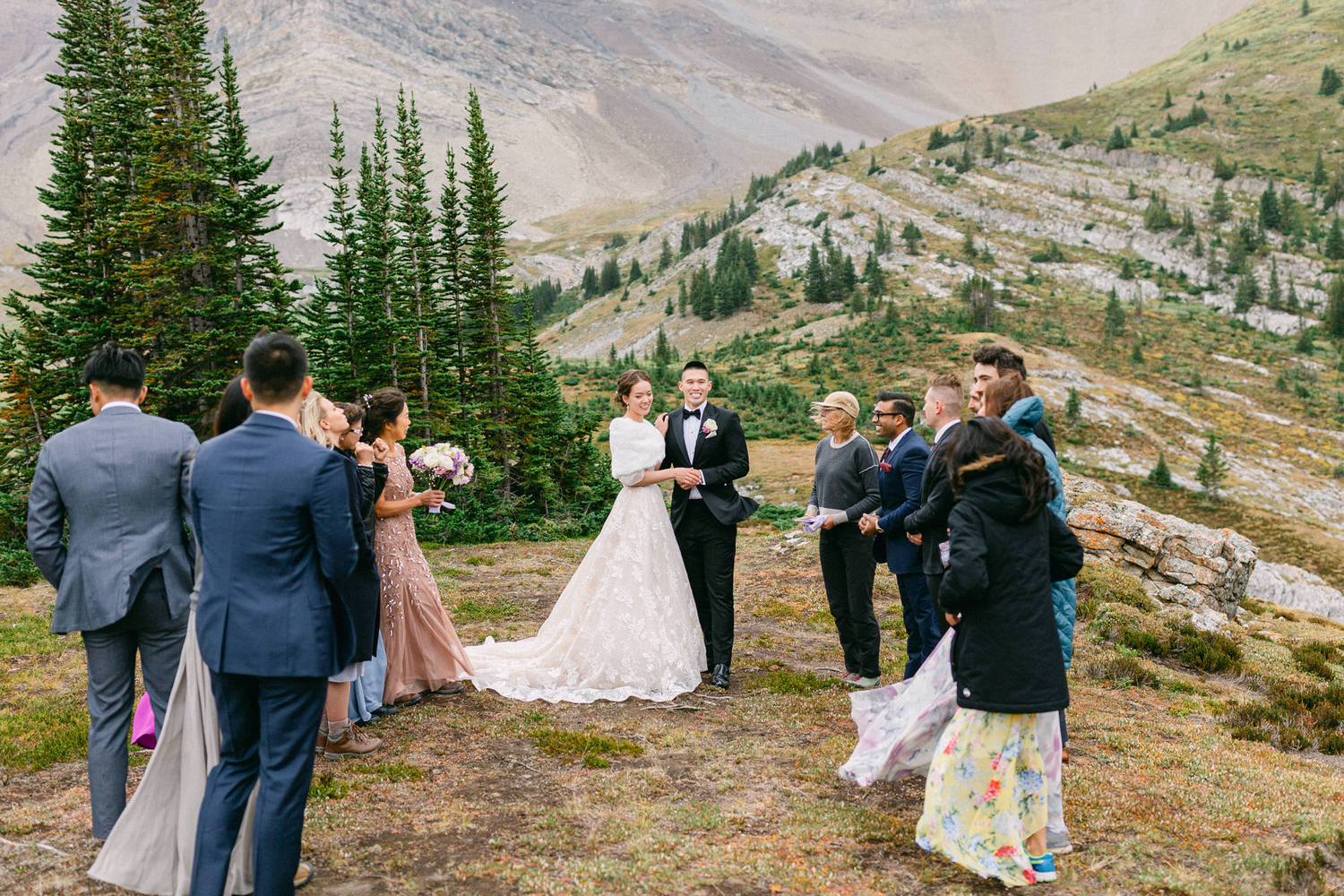 A couple exchanges vows surrounded by friends and family in a beautiful outdoor setting with mountains and trees in the background.