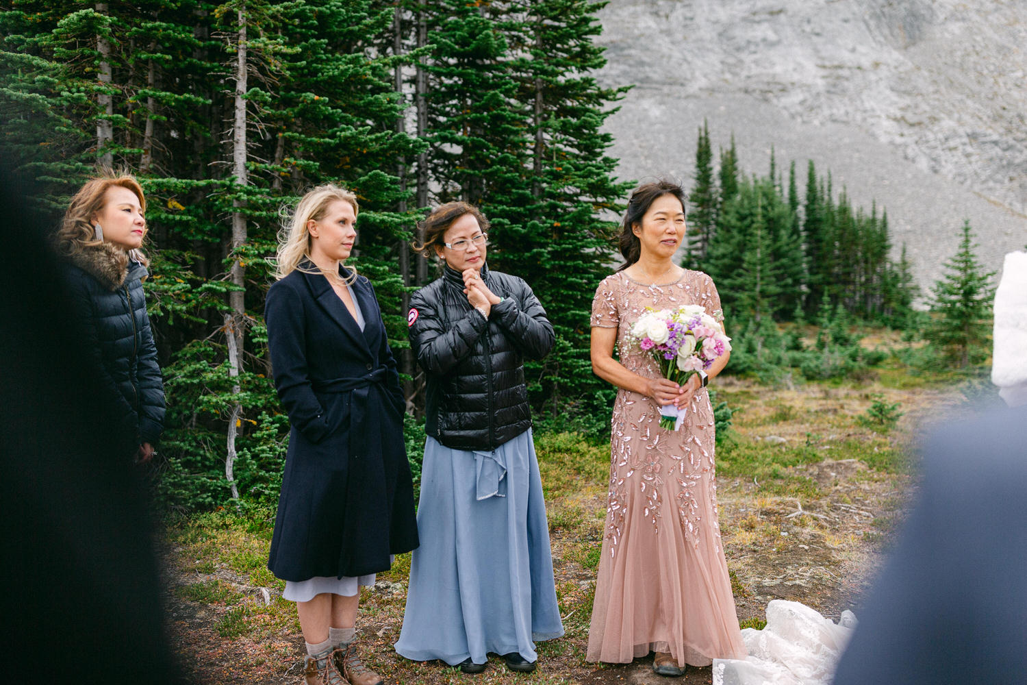 Nature-Inspired Wedding Ceremony::Four women in elegant attire gather outdoors, surrounded by lush greenery, as one holds a bouquet, highlighting a serene wedding moment.