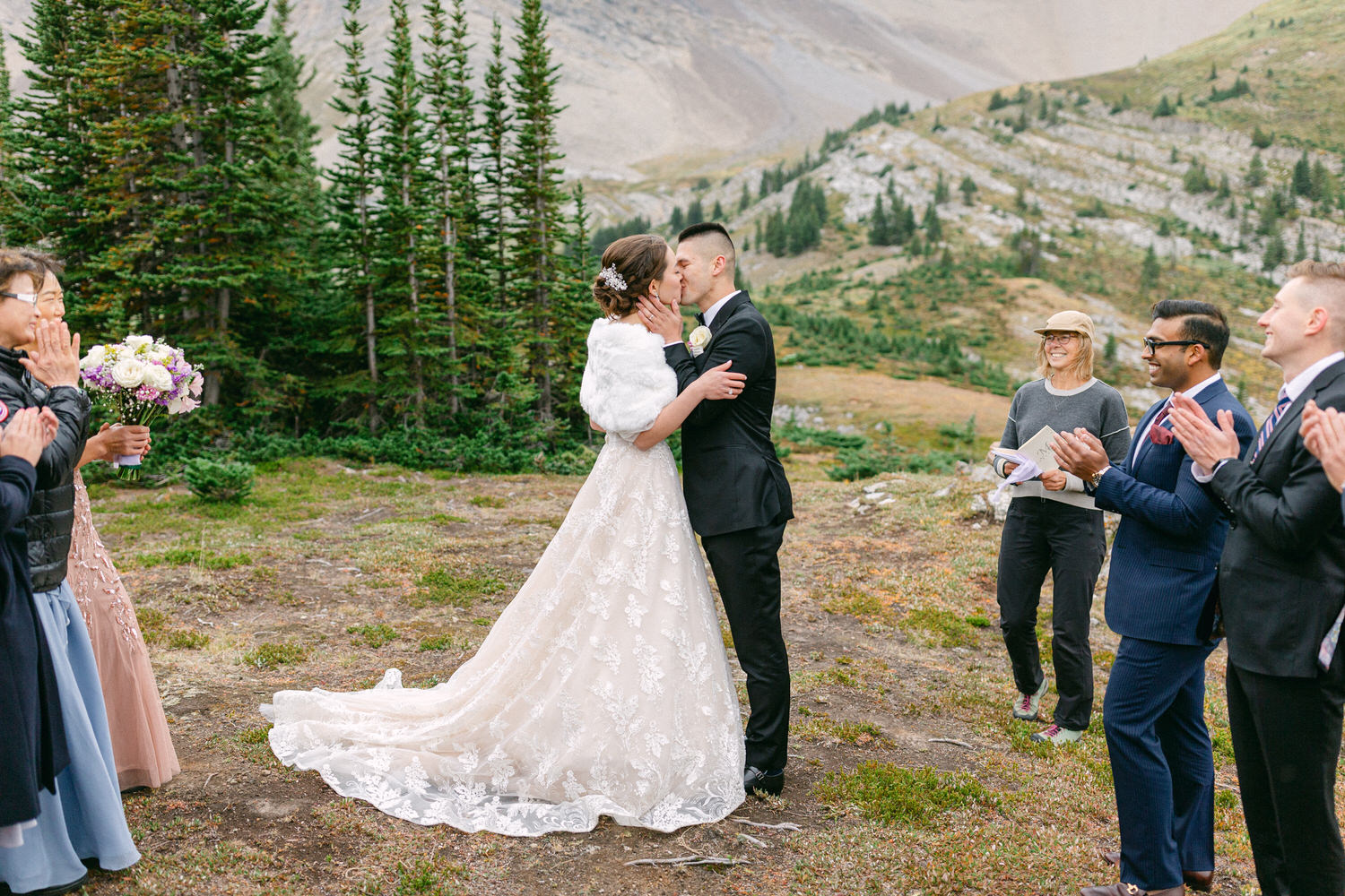 A couple shares a kiss during their outdoor wedding ceremony, surrounded by guests in a scenic mountainous landscape.