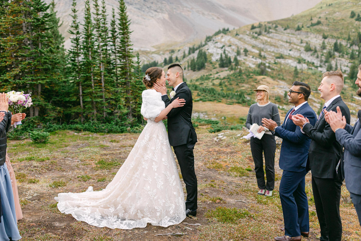 A couple shares a romantic kiss during their outdoor wedding ceremony in a scenic mountain setting, surrounded by friends and family who are applauding.