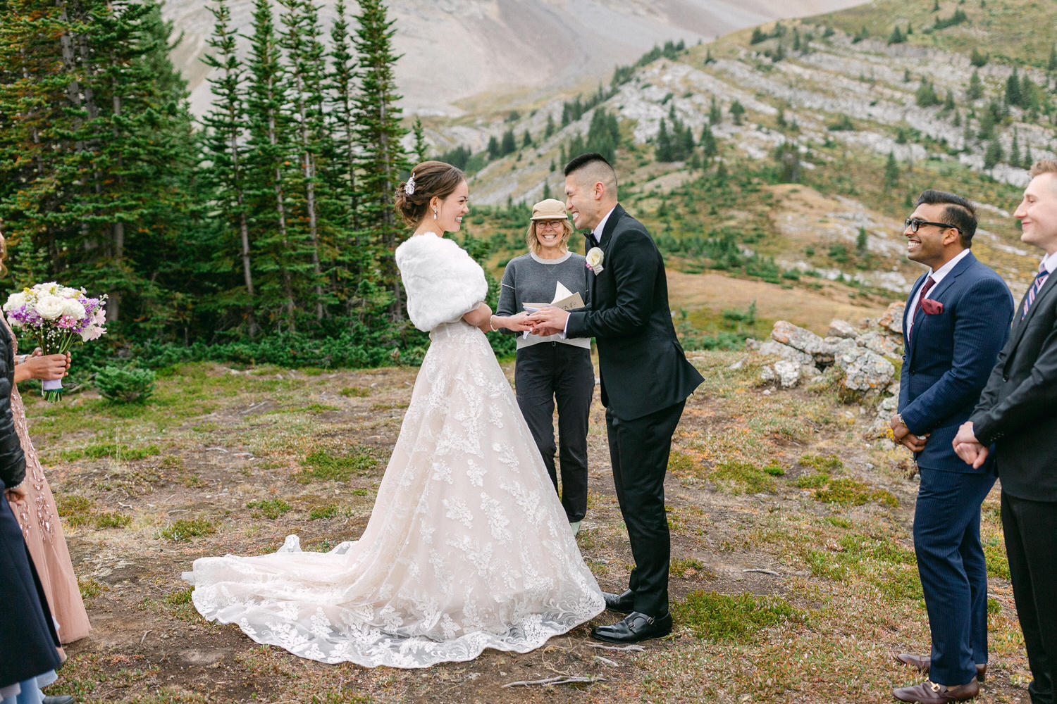 A couple exchanges vows in a scenic outdoor setting, surrounded by friends and a stunning mountain backdrop. The bride wears a beautiful lace gown and cozy shawl, while the groom is in a classic black suit.