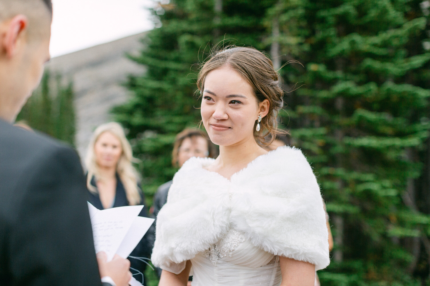 A bride in a white fur shawl smiles during an outdoor wedding ceremony, surrounded by greenery and guests.