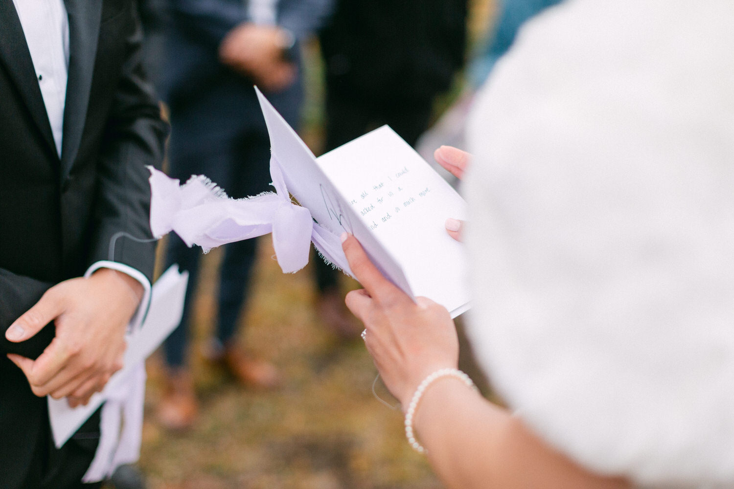 A couple shares heartfelt wedding vows, holding personalized cards while surrounded by guests in a natural outdoor setting.