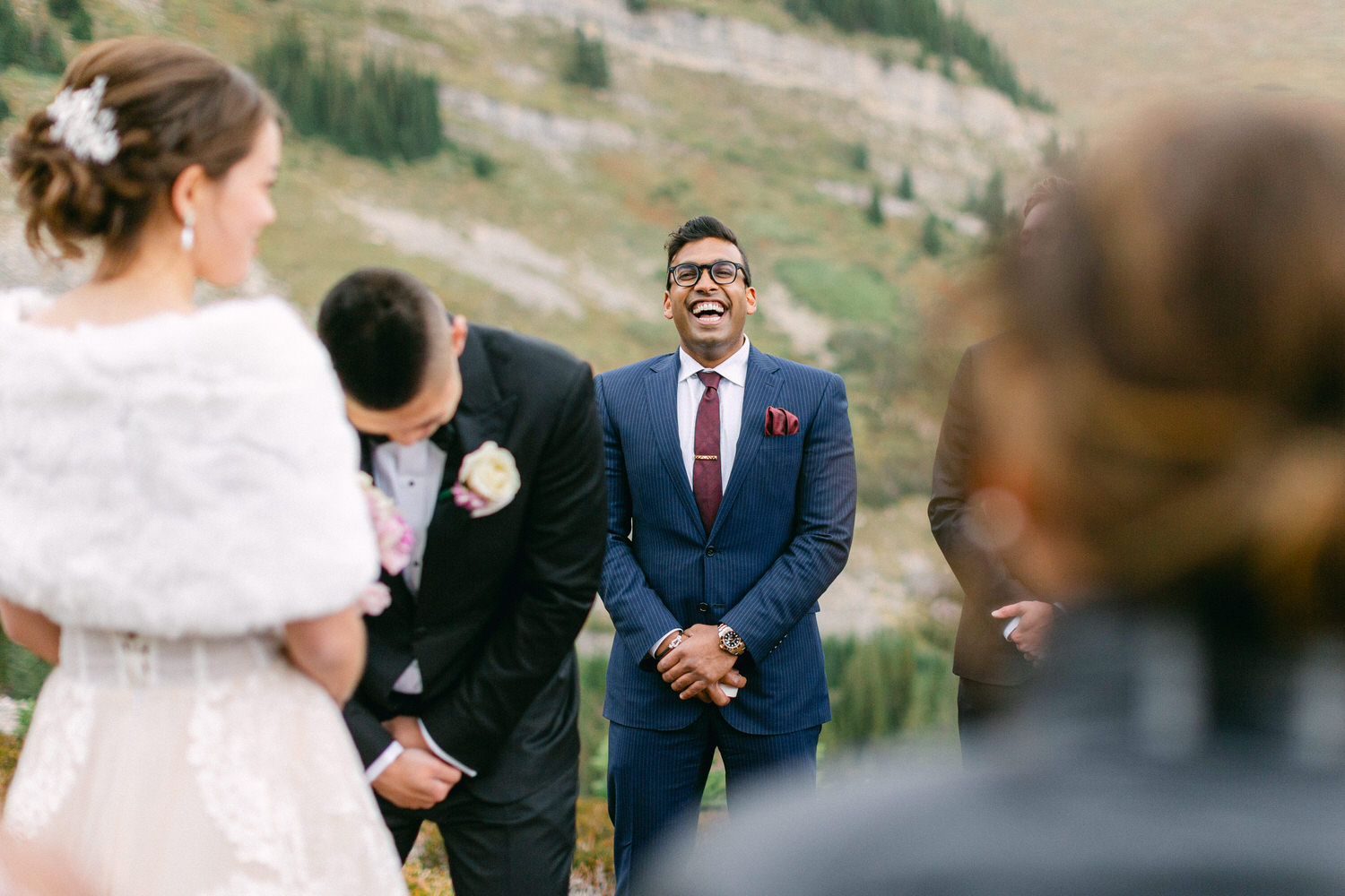 A groom laughs heartily while standing with friends during a serene outdoor wedding ceremony, with the bride partially visible in her elegant attire.