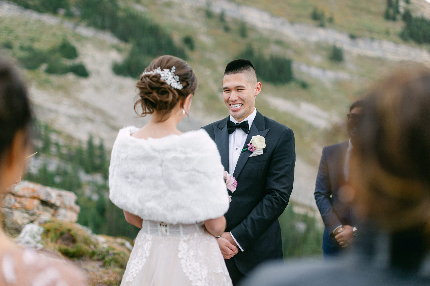 A bride and groom exchange vows in a serene outdoor setting, surrounded by lush greenery and mountains. The bride wears a fur wrap over her wedding dress, while the groom is in a tuxedo, both smiling warmly at each other.