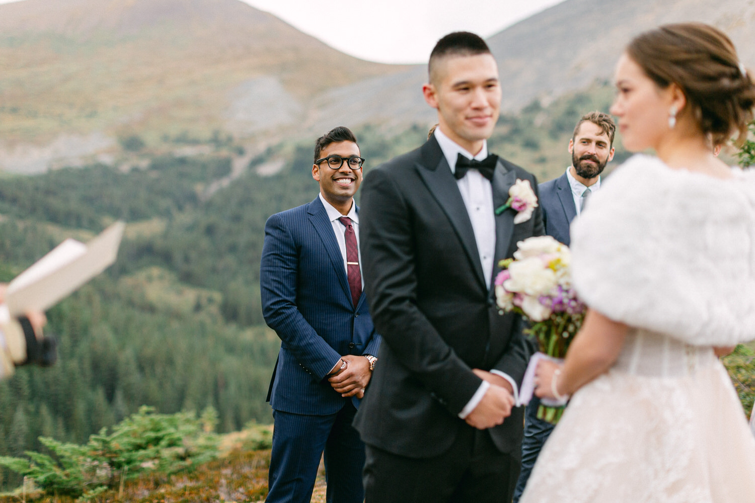 A couple exchanges vows in a scenic mountainous setting, with a smiling guest in the background.