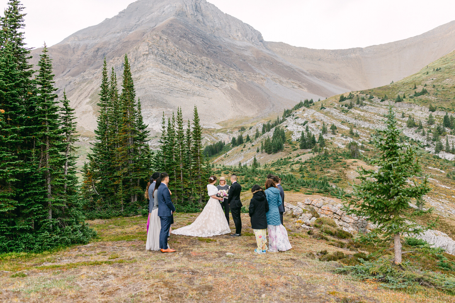 A couple exchanges vows in a serene mountain setting, surrounded by guests and evergreen trees.