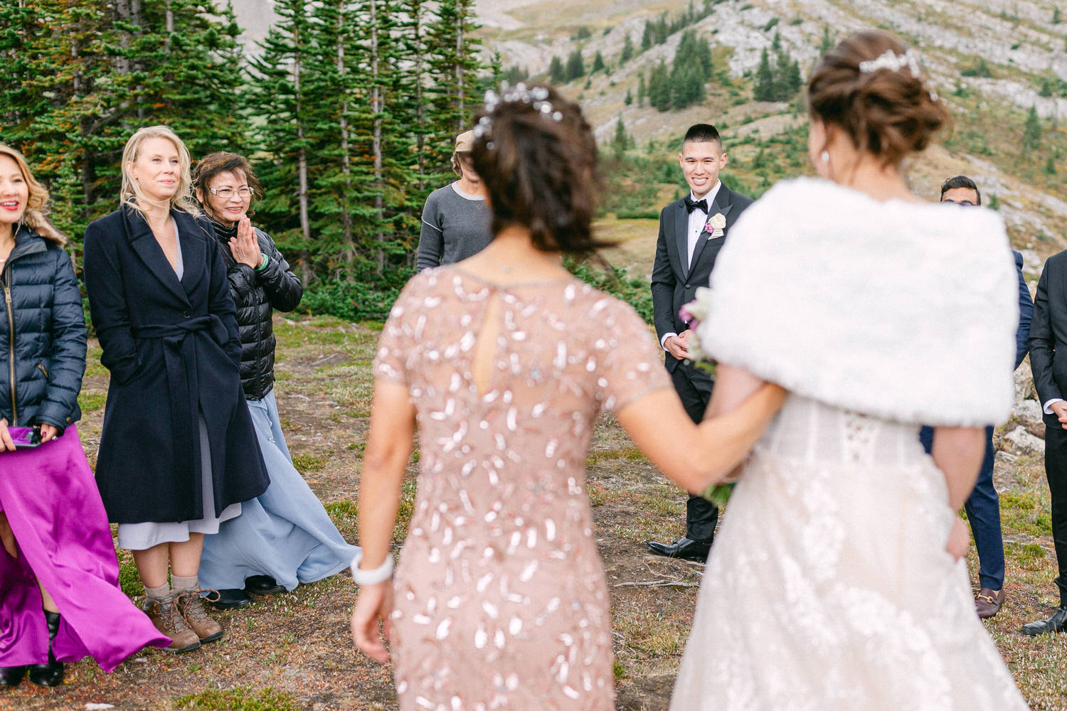 A couple stands together in an outdoor ceremony, surrounded by guests in formal attire, with a scenic mountainous backdrop.