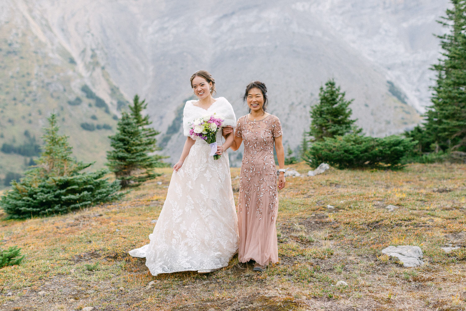 A bride in a white lace gown and her companion in a peach dress, standing together in a scenic mountainous landscape, with green trees and a rocky backdrop.