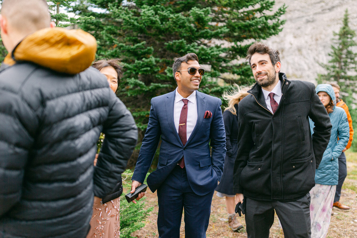 A group of well-dressed individuals laughs and converses in a forest setting, with trees in the background and a mix of formal and casual attire.